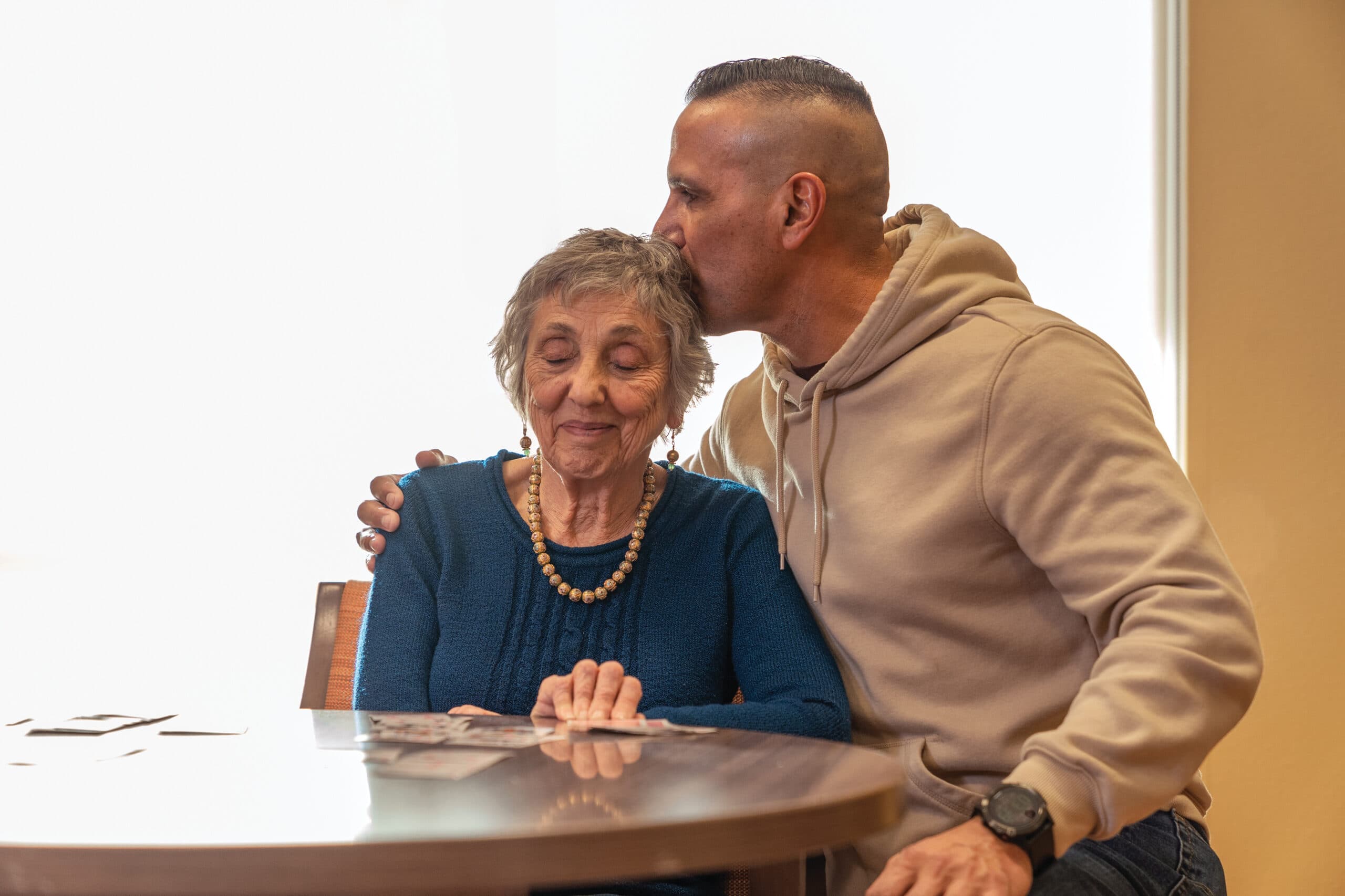 A man kissing the forehead of an elderly woman as she sits at a table playing cards.