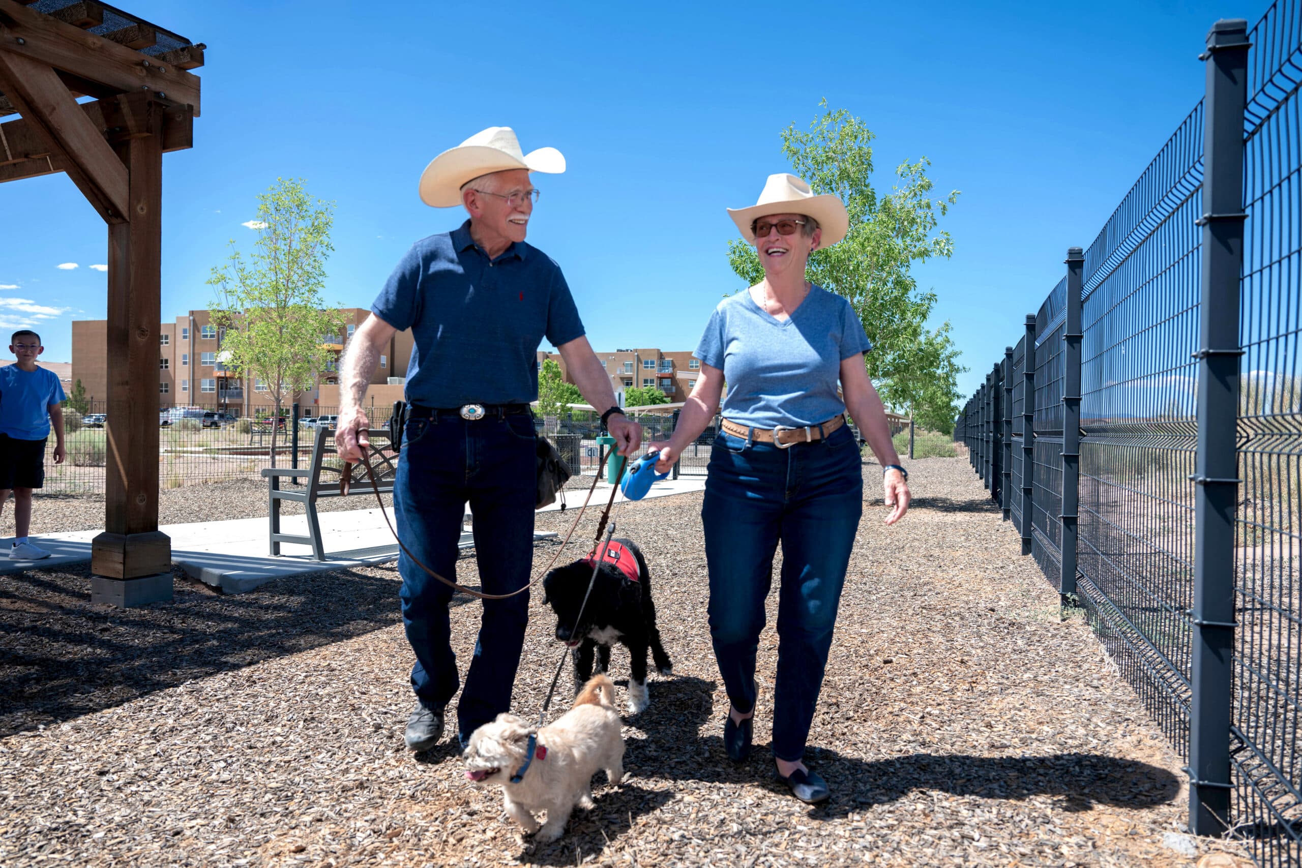 A man and woman walking two leashed dogs along a gravel path beside a fence under a clear blue sky.