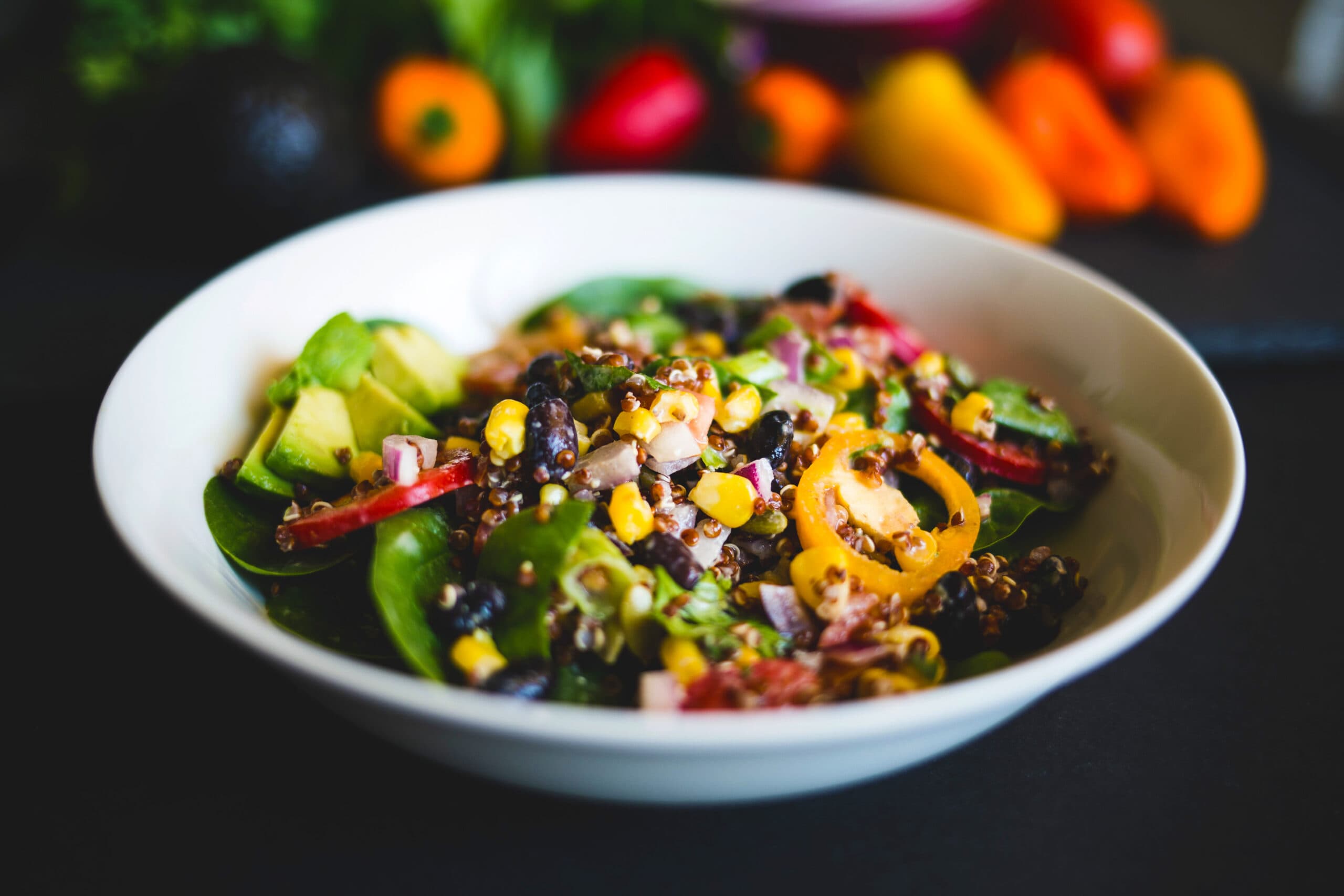 Quinoa salad with corn, black beans, diced avocado, and mixed greens in a white bowl with blurred vegetables in the background.