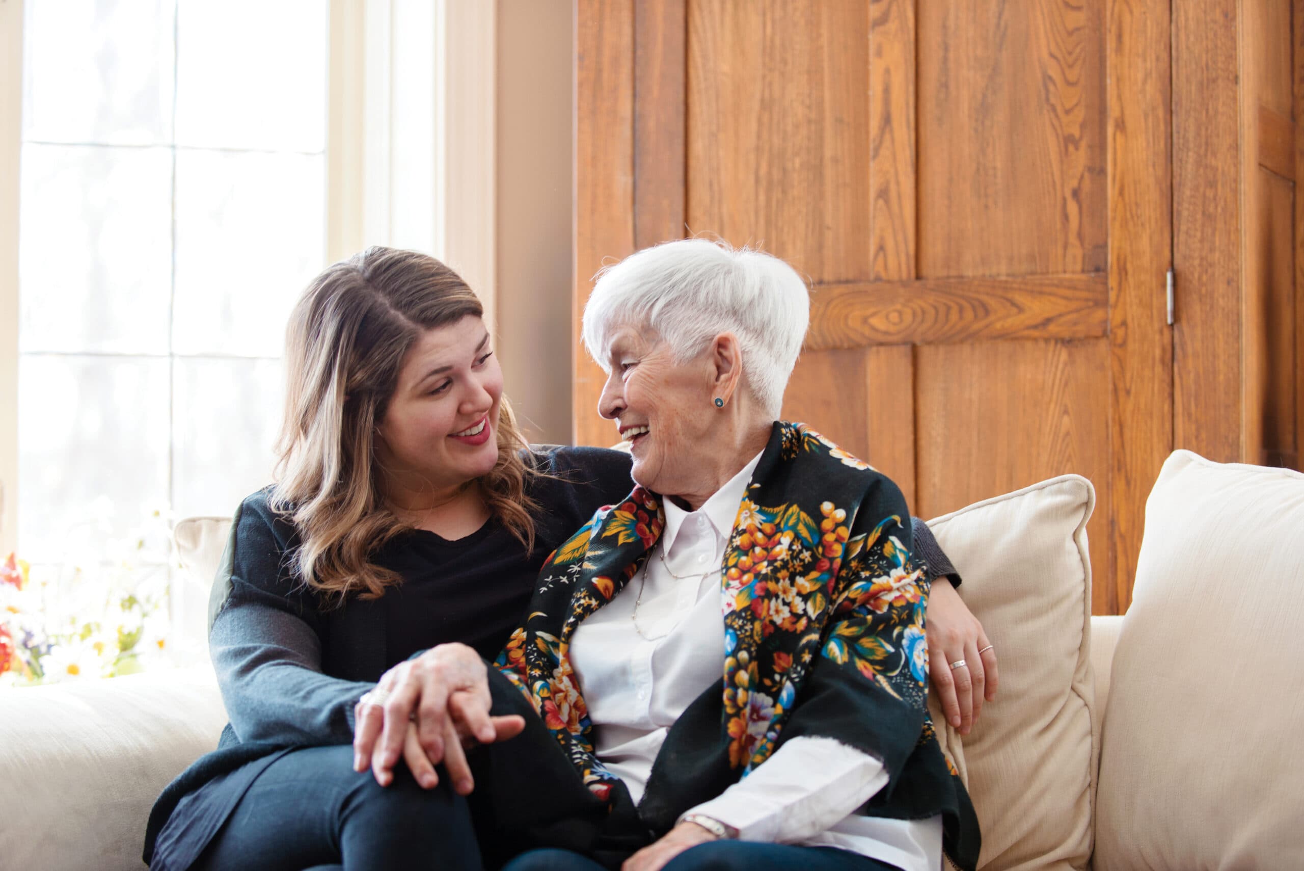 A younger woman and an elderly woman sitting on a couch, smiling and holding each other's arms.