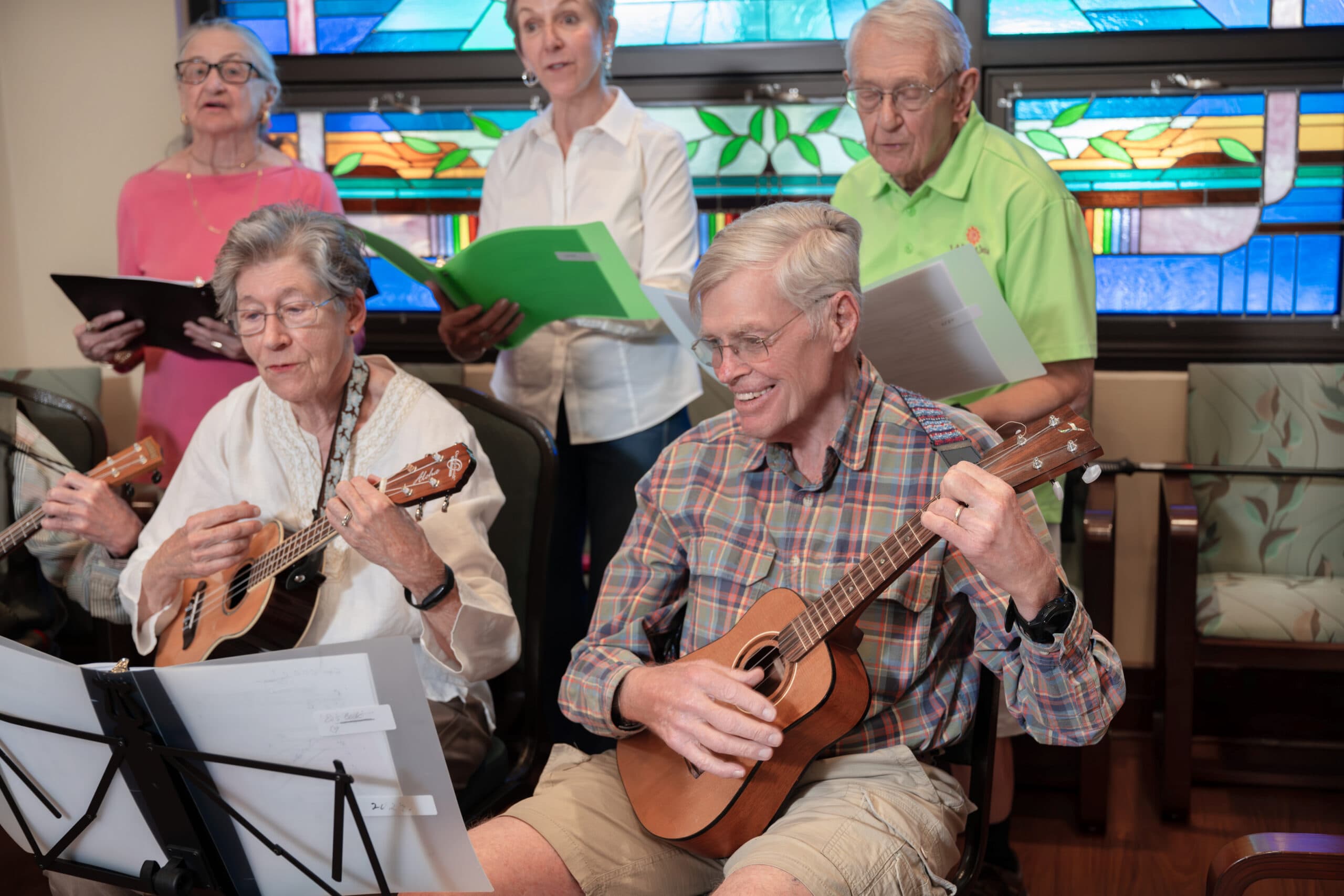 Older adults play ukuleles and sing together by colorful stained-glass windows.